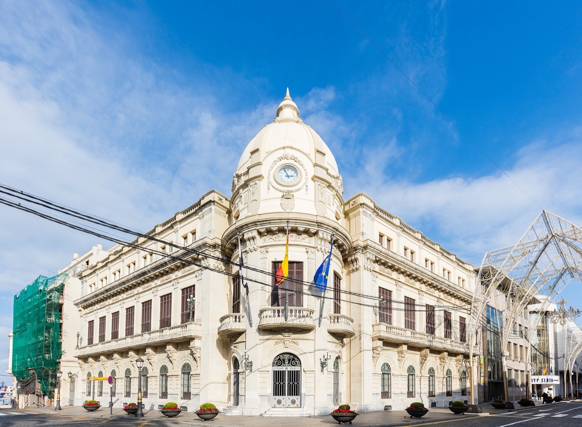 Fachada blanca del Palacio de la Asamblea de Ceuta, con su cúpula y su reloj, y las banderas en el balcón central