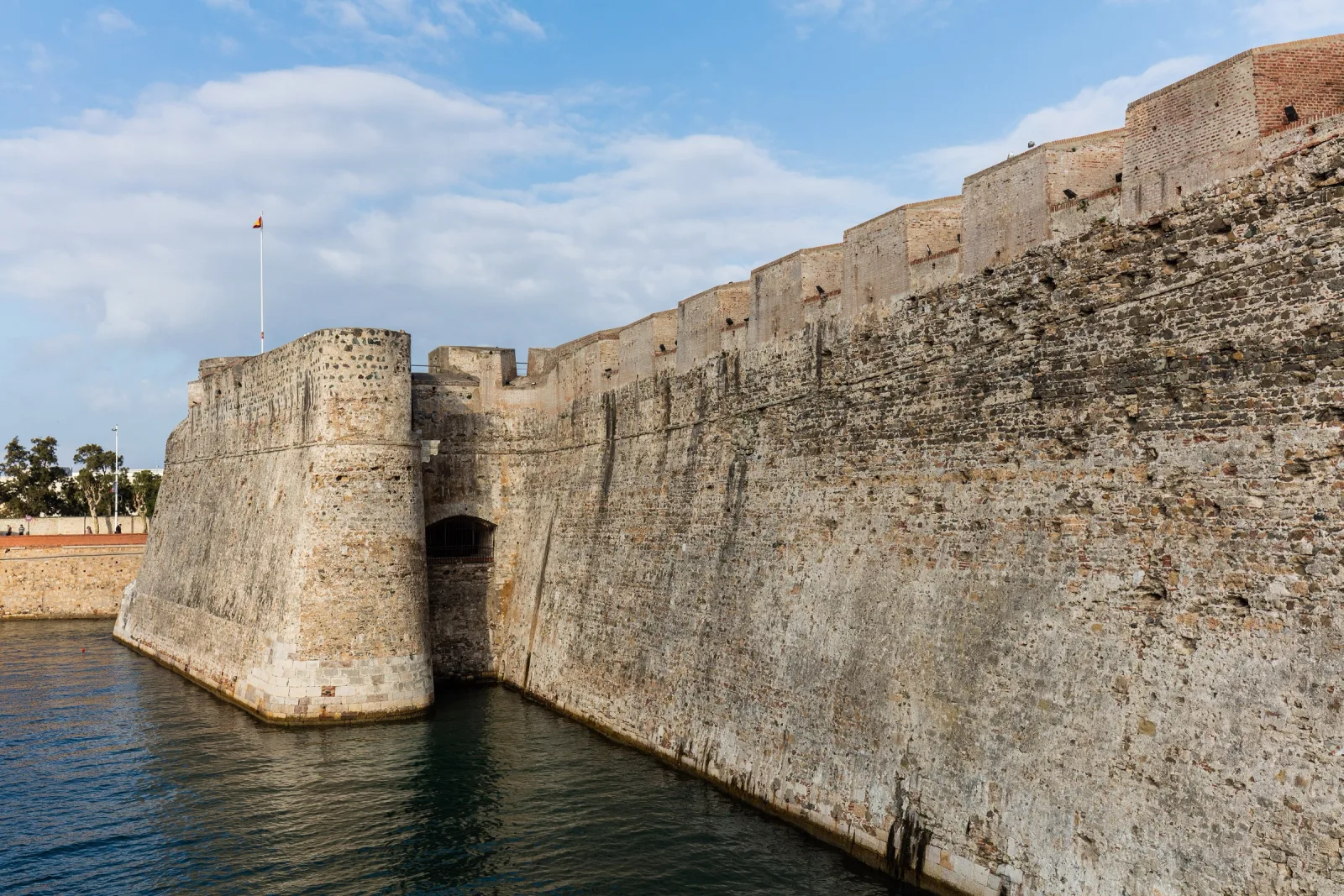 Altos muros de piedra de las Murallas Reales de Ceuta junto al foso inundado de agua, con un baluarte en primer plano y cielo azul con nubes.
