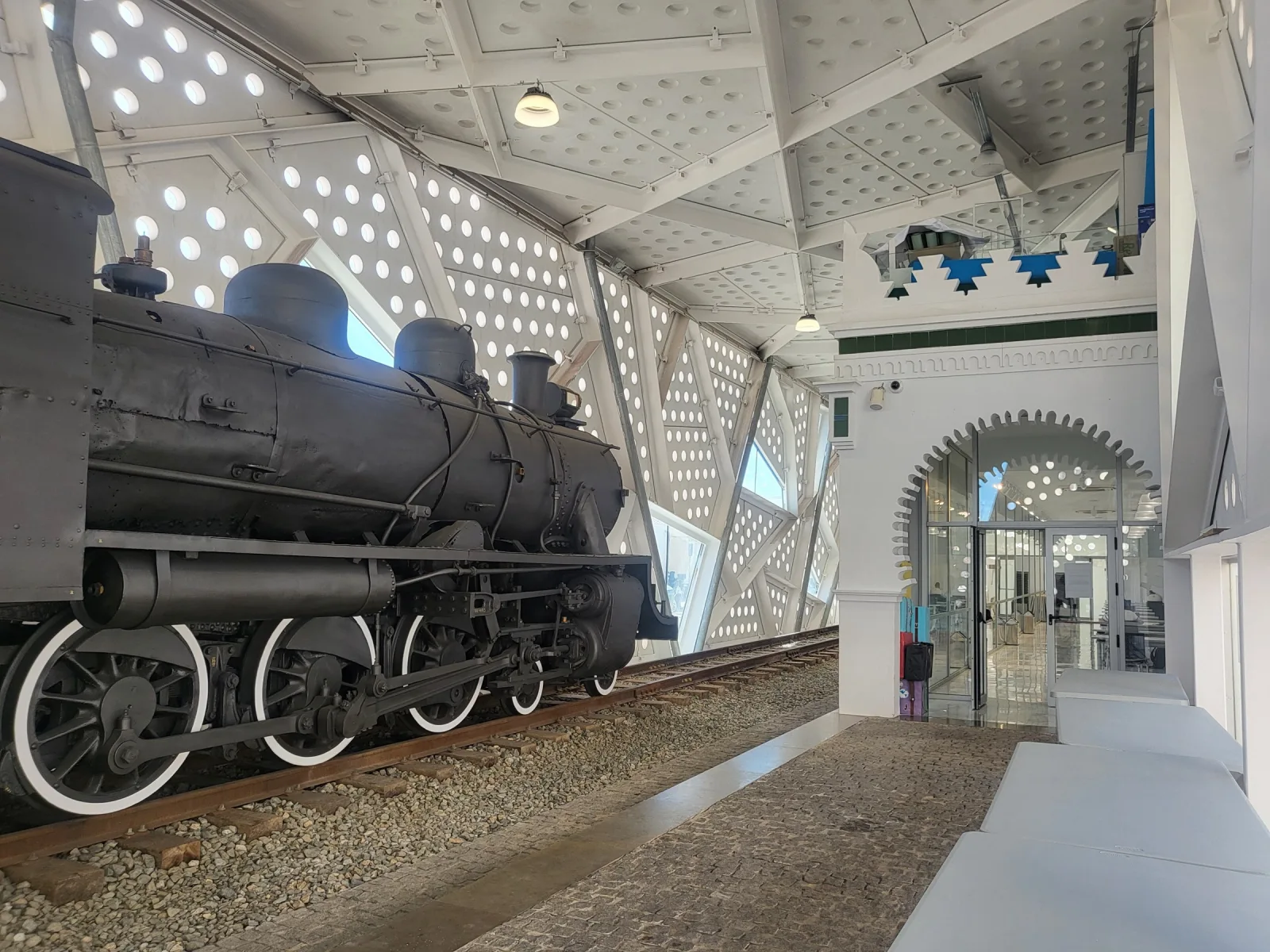 Locomotora de vapor negra sobre un tramo de vía, bajo una cubierta moderna de paneles blancos perforados, junto a la entrada en arco de la antigua estación de Ceuta.