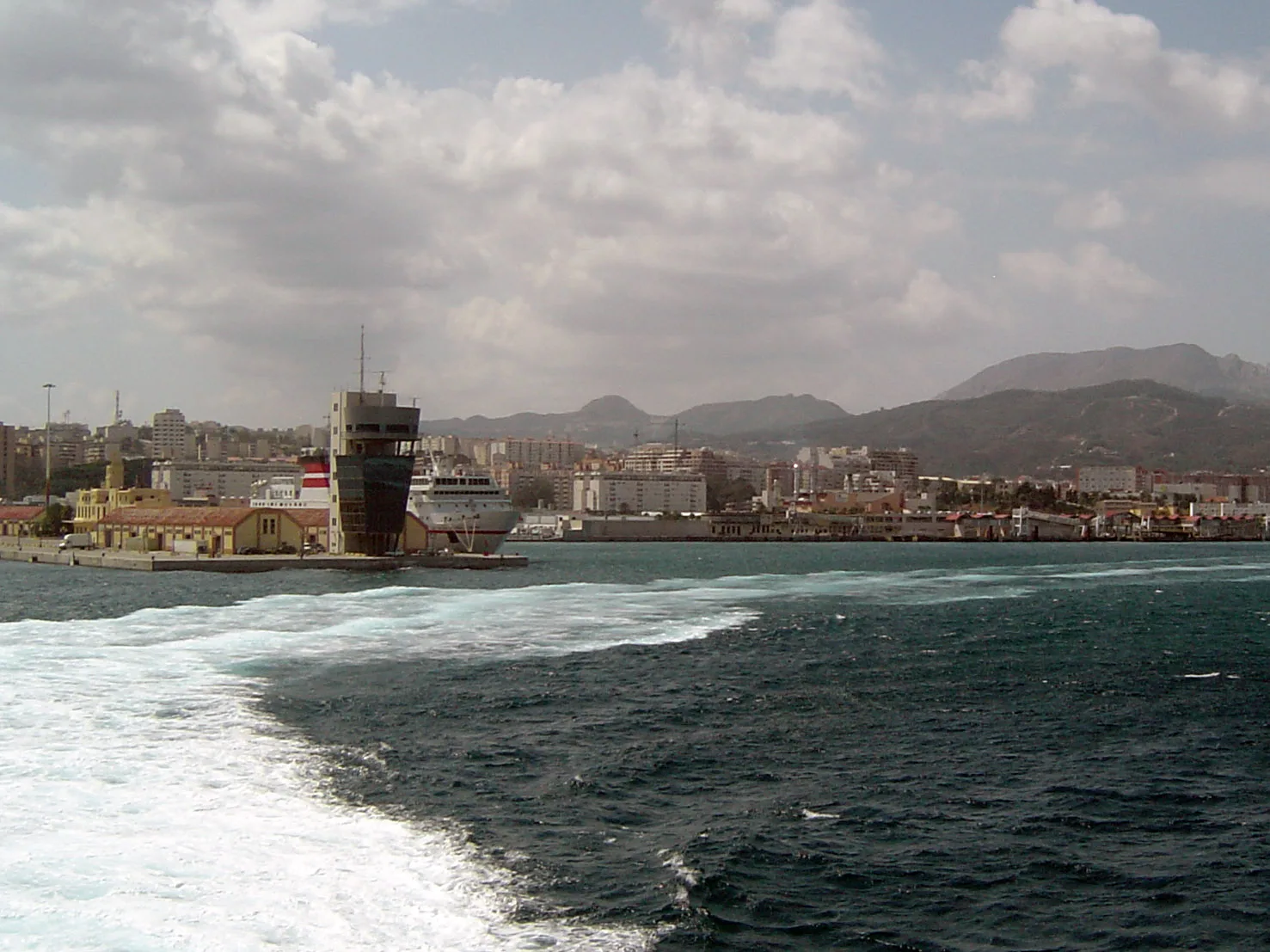 El puerto de Ceuta visto desde la popa de un barco que se aleja, con la estela blanca en primer plano, la torre de control, un ferry atracado y los montes detrás.