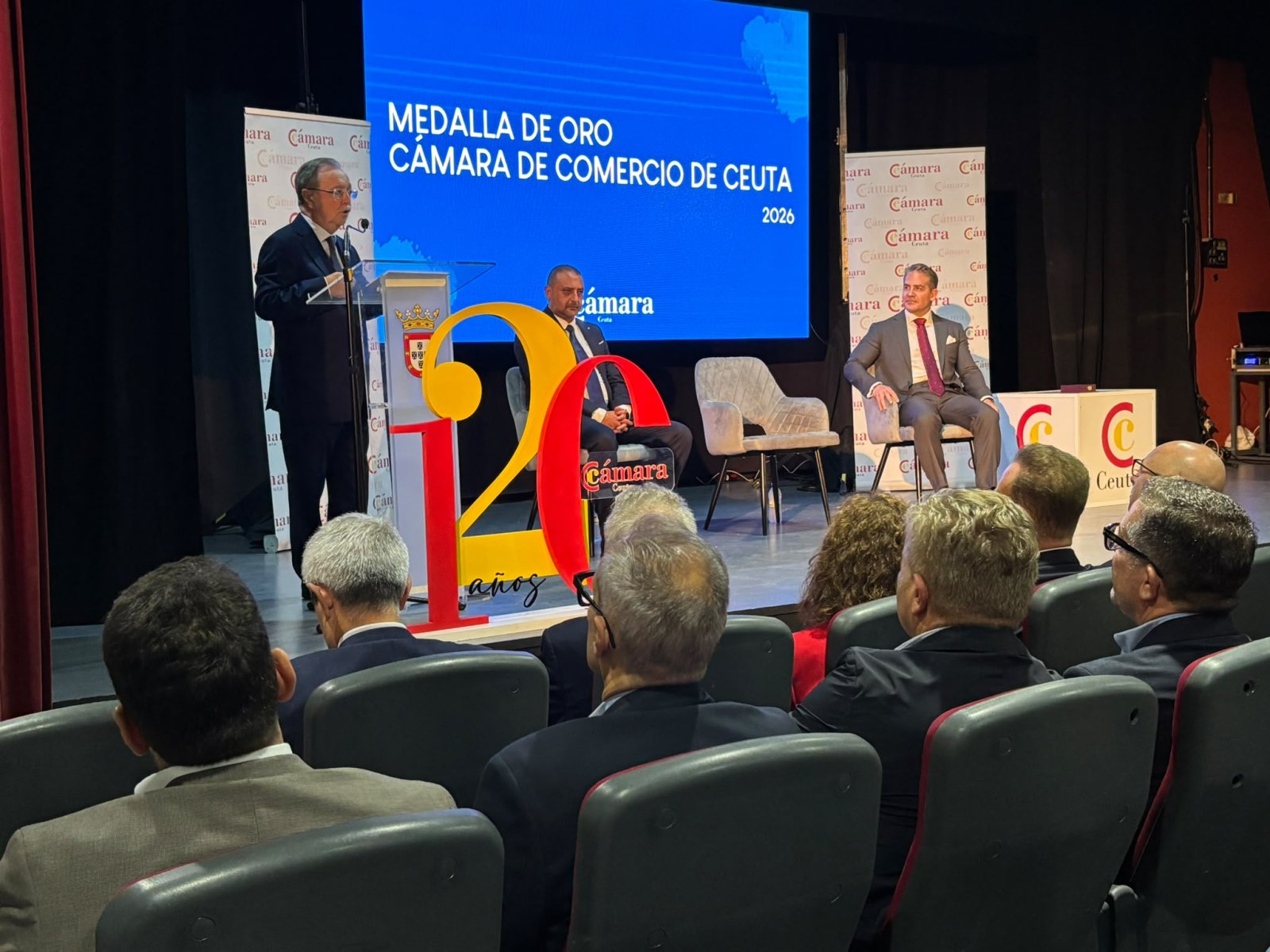Un hombre habla en un atril ante un público sentado, con una pantalla azul que dice Medalla de Oro Cámara de Comercio de Ceuta 2026.
