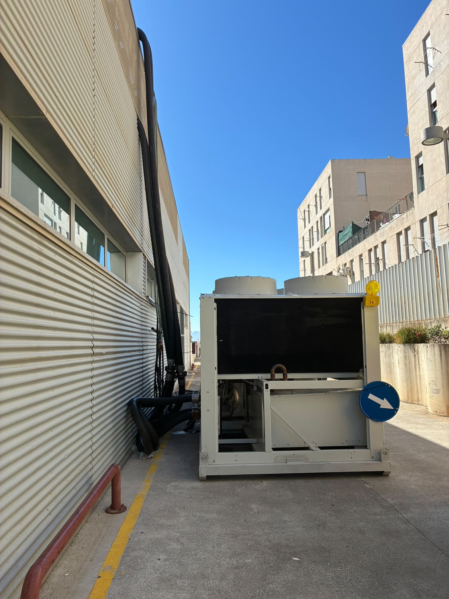 Una gran unidad de refrigeración industrial con ventiladores en la parte superior, junto a gruesos tubos negros adosados a la pared de chapa de un edificio, en