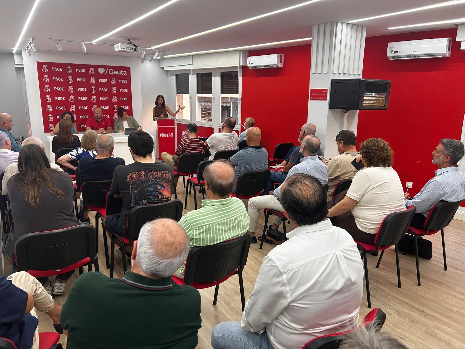 Una mujer habla desde un atril ante un público sentado en sillas, en una sala con paredes rojas y un cartel del PSOE de Ceuta.