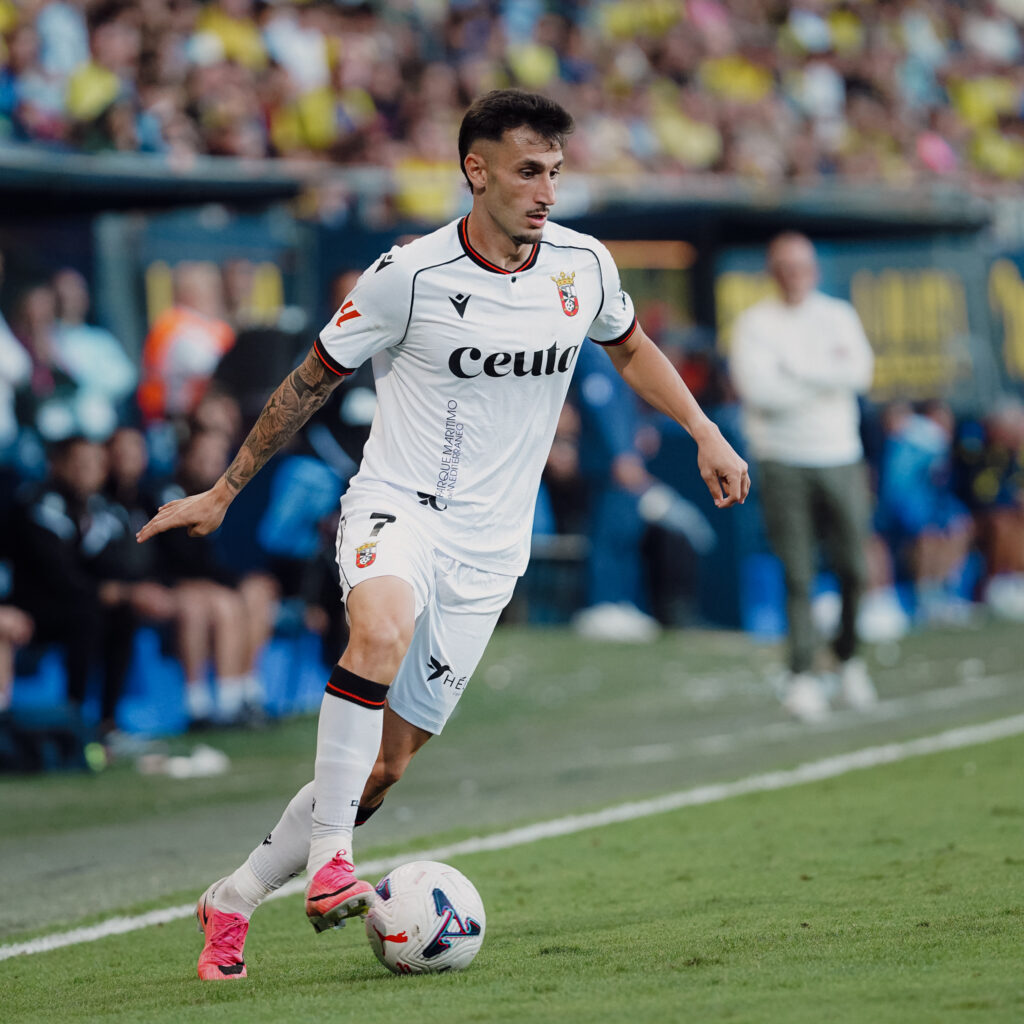 Un futbolista con camiseta blanca y el texto Ceuta conduce el balón sobre el césped, con público y banquillo al fondo.