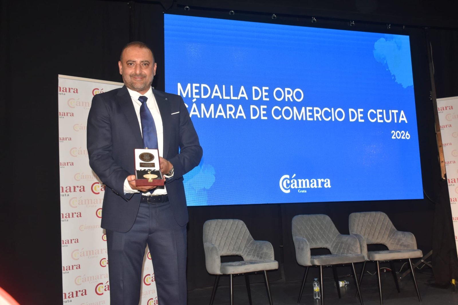 Un hombre trajeado sostiene una medalla ante una pantalla azul que dice Medalla de Oro Cámara de Comercio de Ceuta 2026.