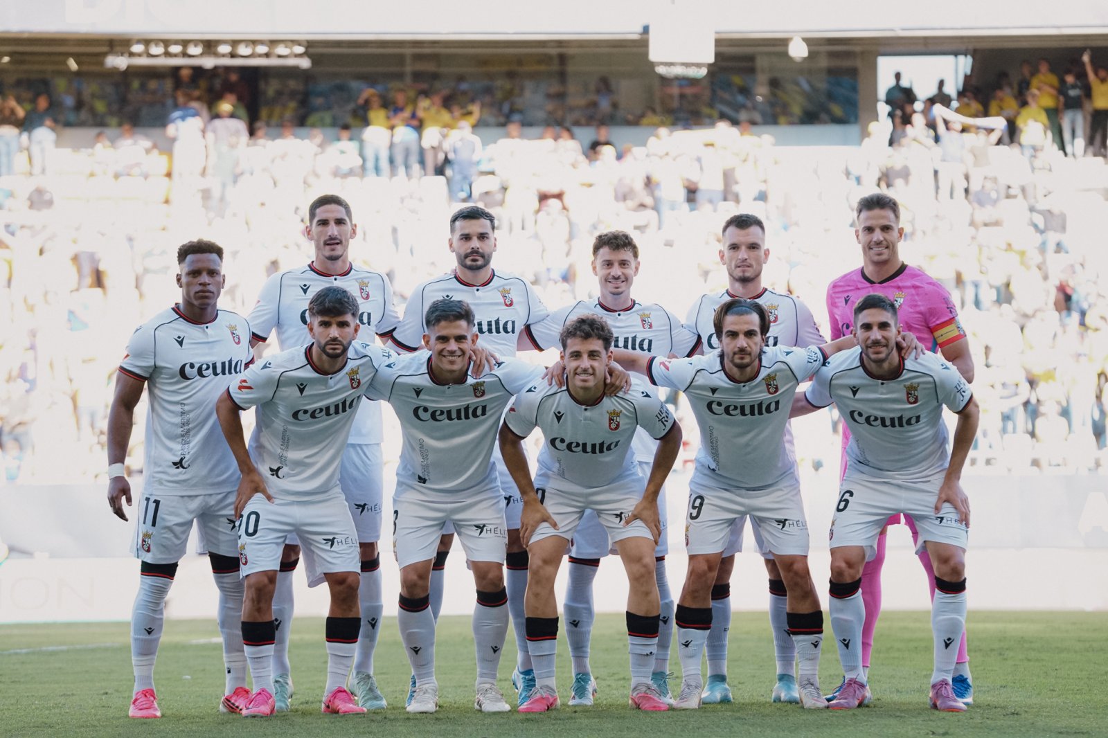 Once jugadores de fútbol con equipación blanca posan en grupo sobre el césped, con un portero de rosa y público en las gradas al fondo.
