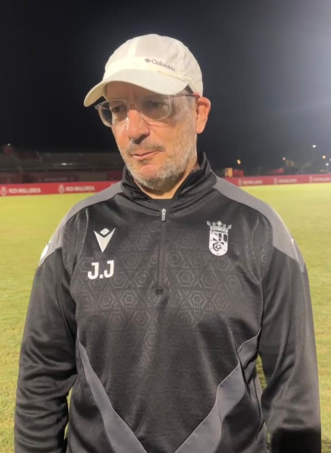 Un hombre con gorra clara y gafas, con sudadera negra de entrenador, de noche en un campo de fútbol.