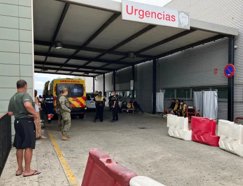 Entrada de Urgencias con una ambulancia, personal sanitario y militares junto a personas sentadas y barreras.