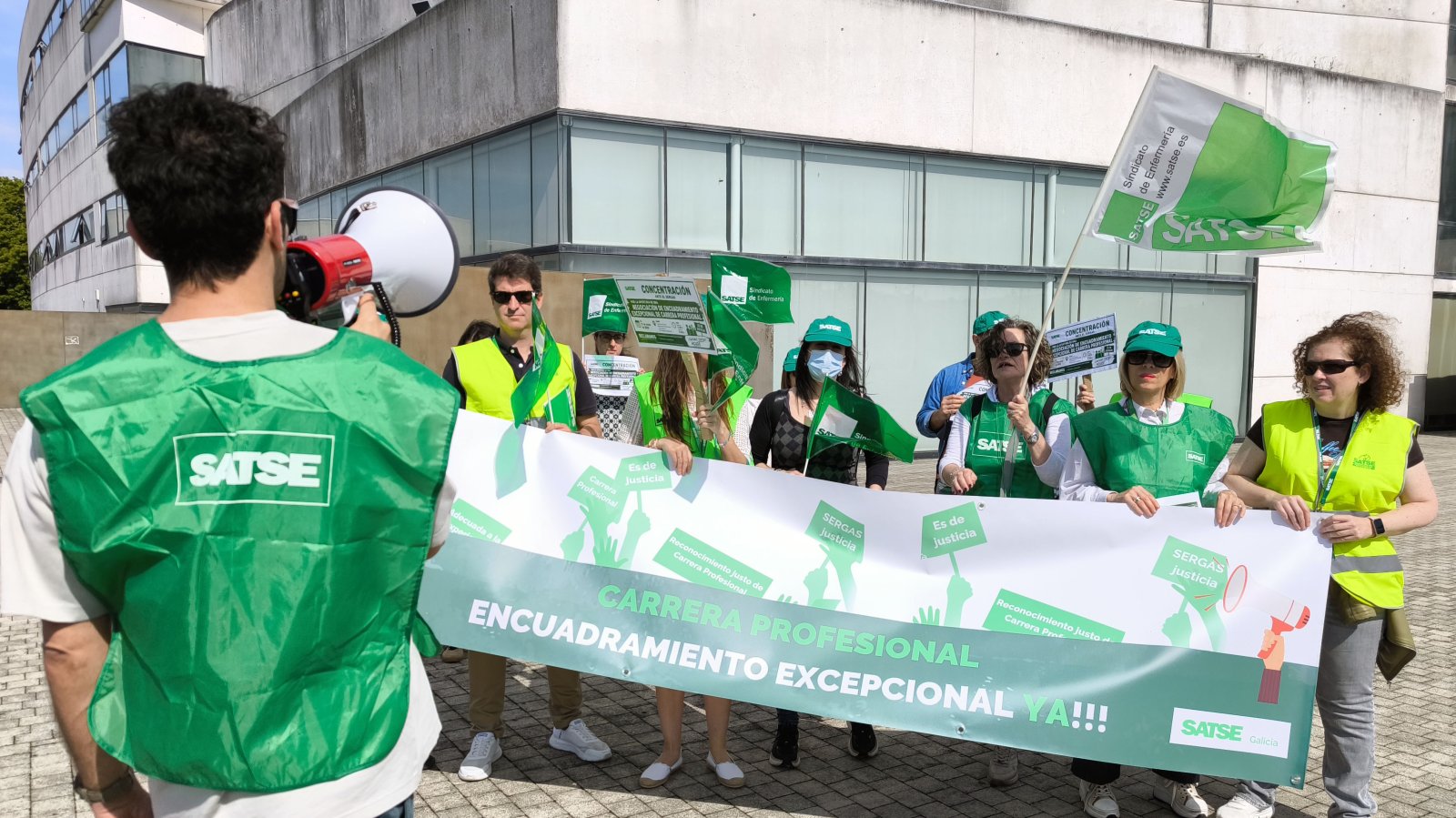 Un grupo de personas con chalecos verdes y banderas de SATSE sostiene una pancarta reivindicativa ante un edificio.