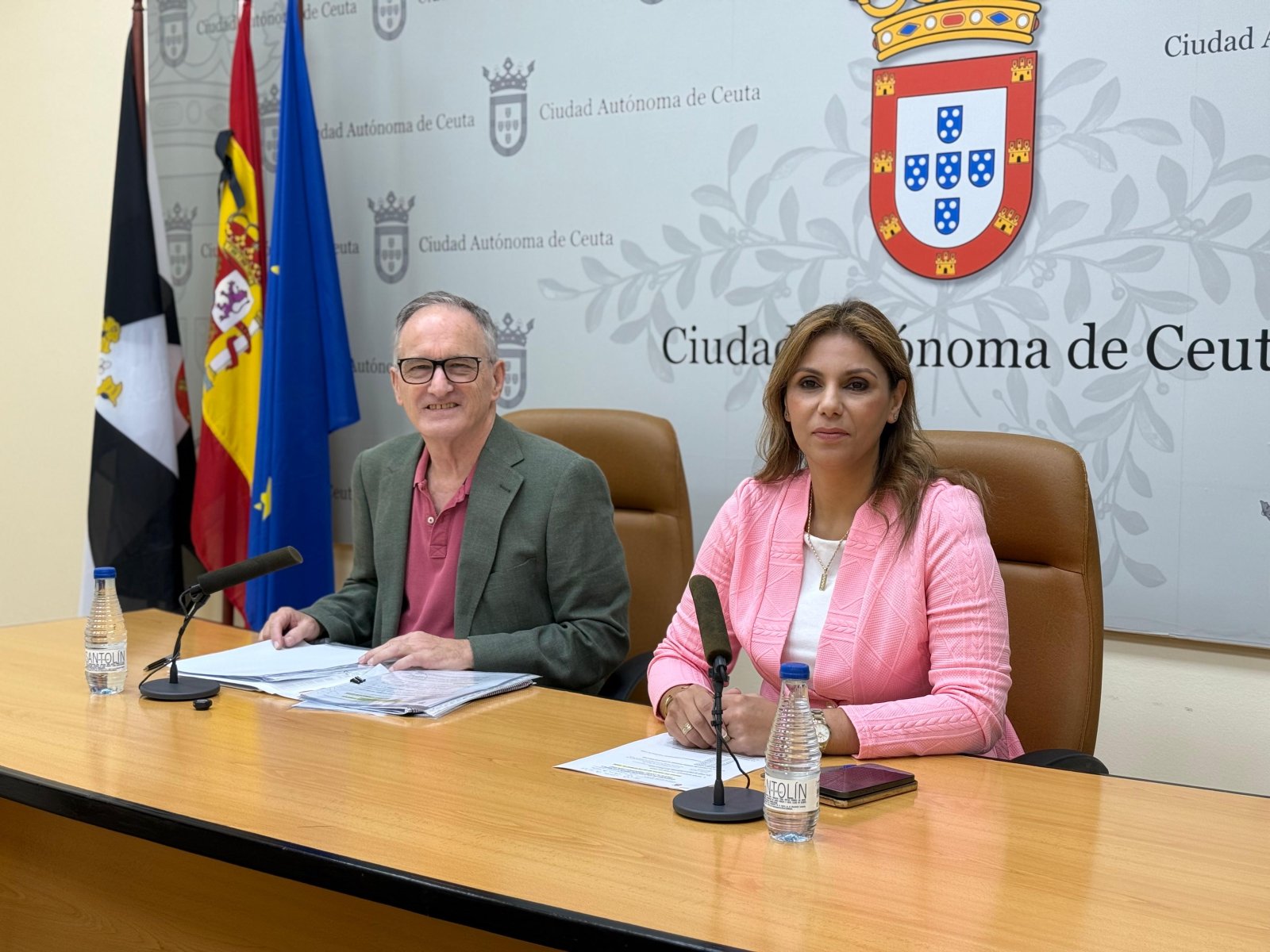 Un hombre con gafas y una mujer con chaqueta rosa sentados ante una mesa con micrófonos y documentos, con banderas y el escudo de Ceuta al fondo.